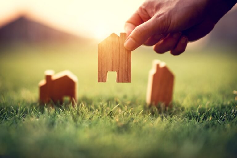 hand-of-woman-choosing-mini-wood-house-model-on-green-grass-planning-buy-real-estate-eco-house-icon-concept-min-768×513